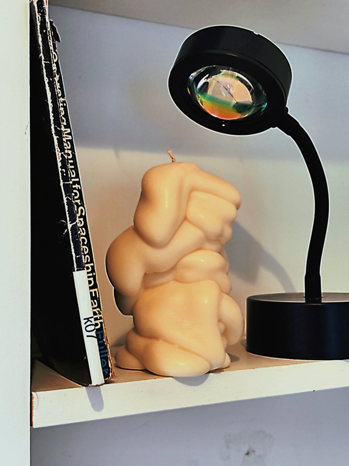 Drape sculptural candle by Post Objects on a white shelf beside a black desk lamp and book