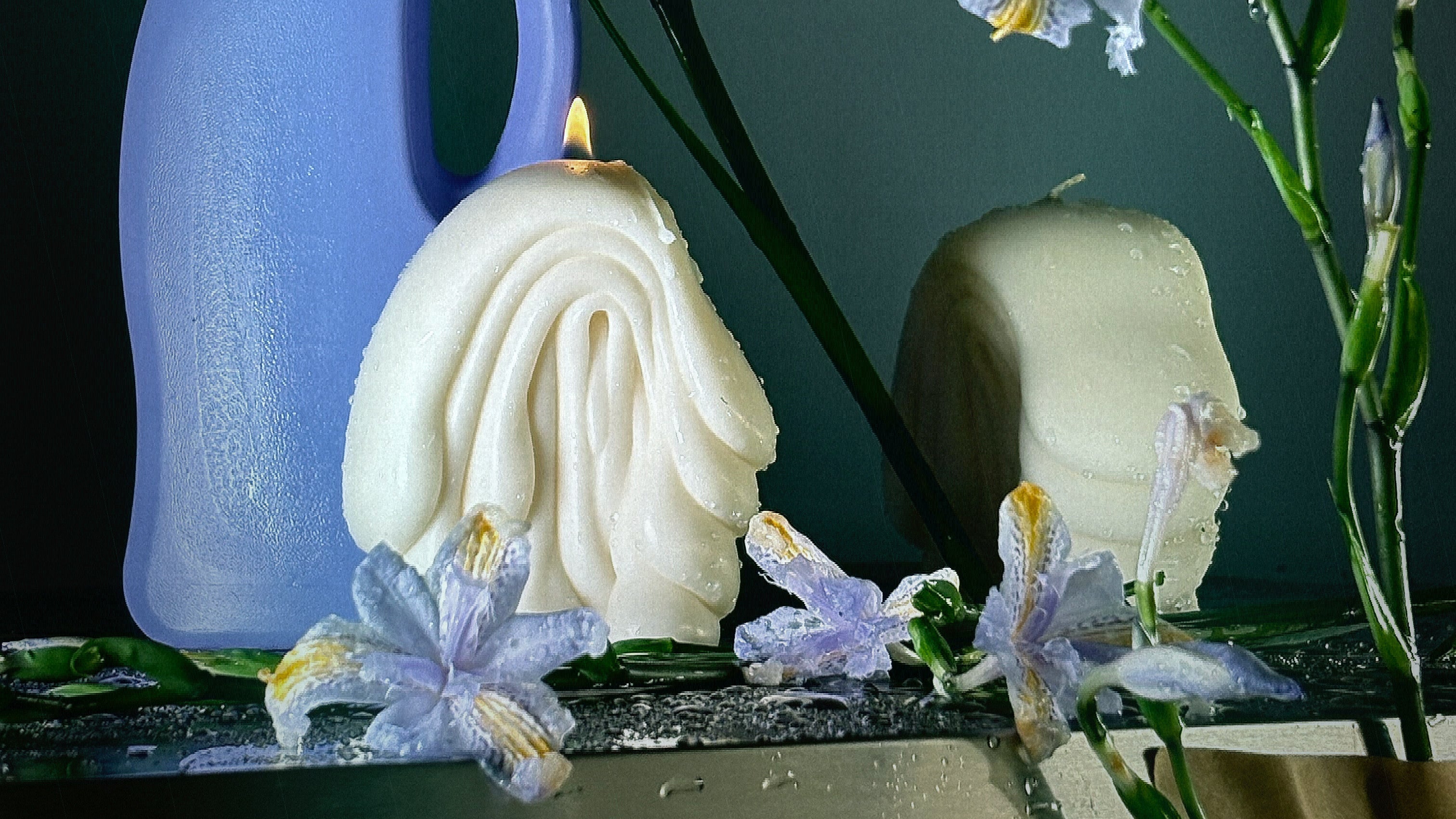A styled product still life featuring a lavender-blue plastic bottle, a sculpted white Post Objects Fold abstract candle, and light purple iris flowers on a dark reflective surface against a dark green background.