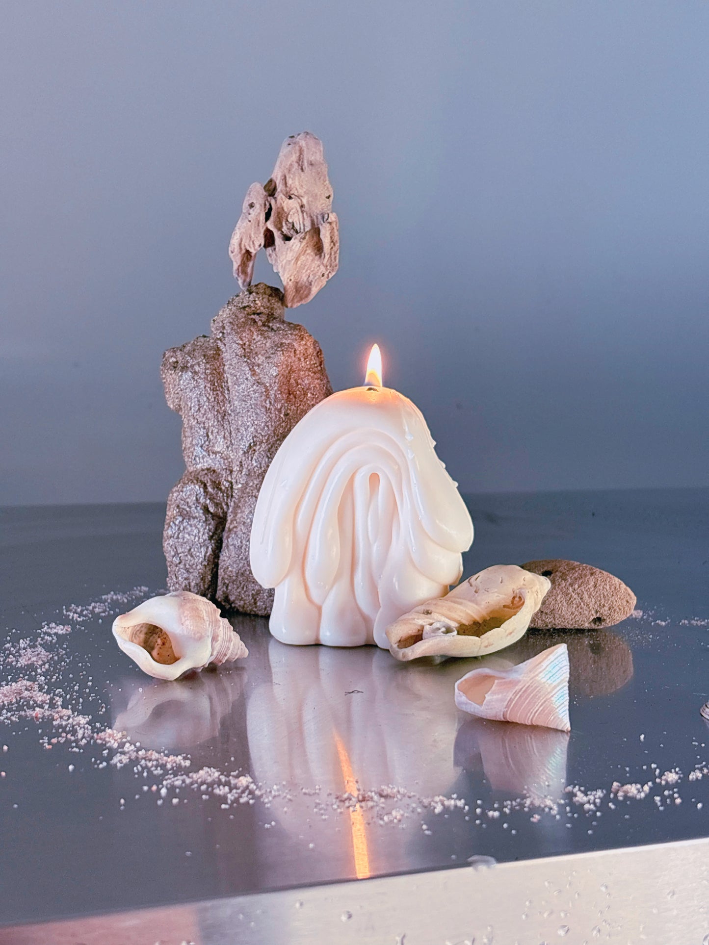 A Post Objects sculptural candle shaped like an organic folded drippy blob, lit with a small flame, displayed on a reflective surface with driftwood, shells, and sand against a soft blue-gray background.