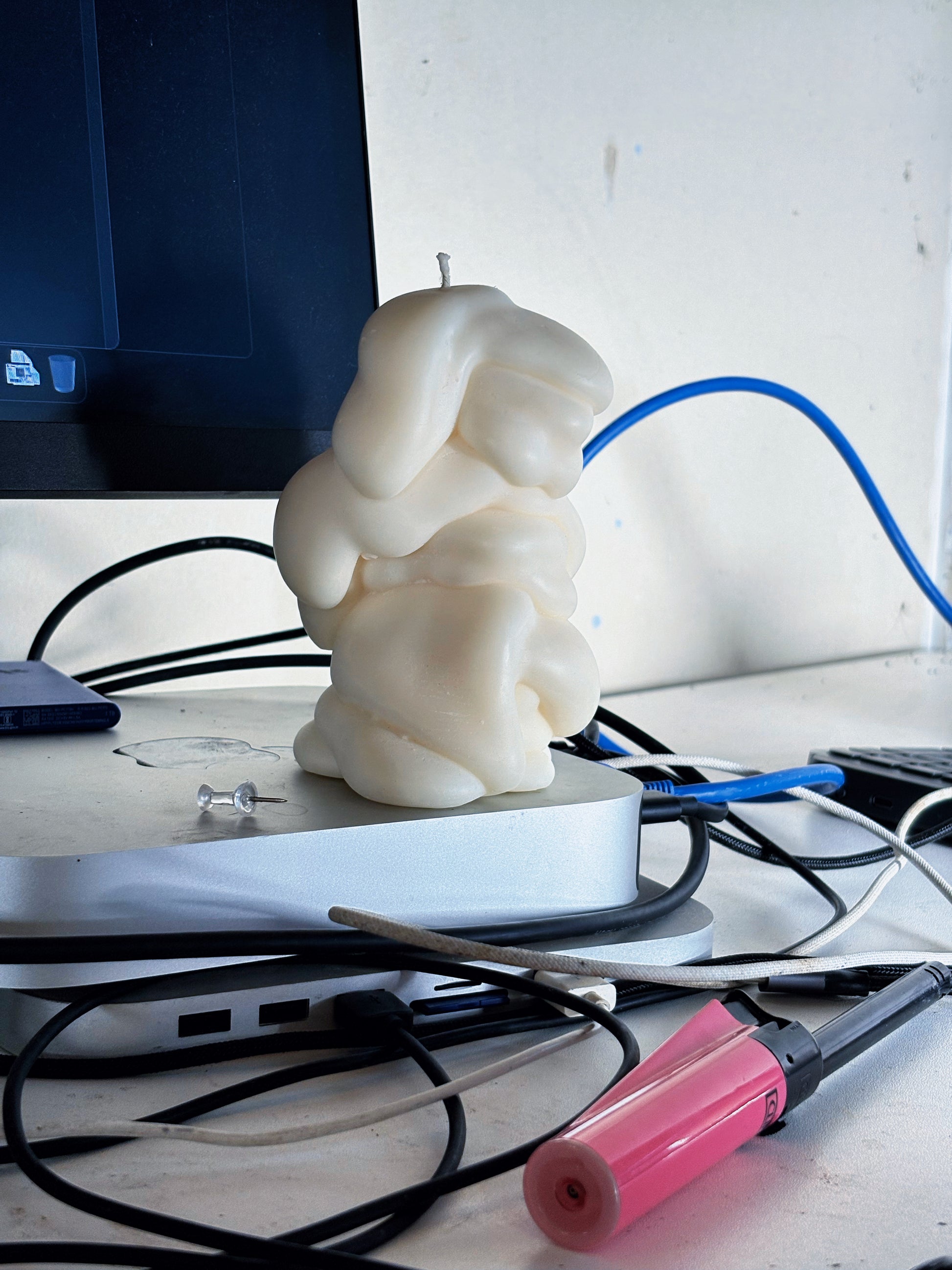 A post objects sculptural cream-colored candle shaped like a an abstract draped blob sits on a desk cluttered with computer cables, a monitor, and a lighter