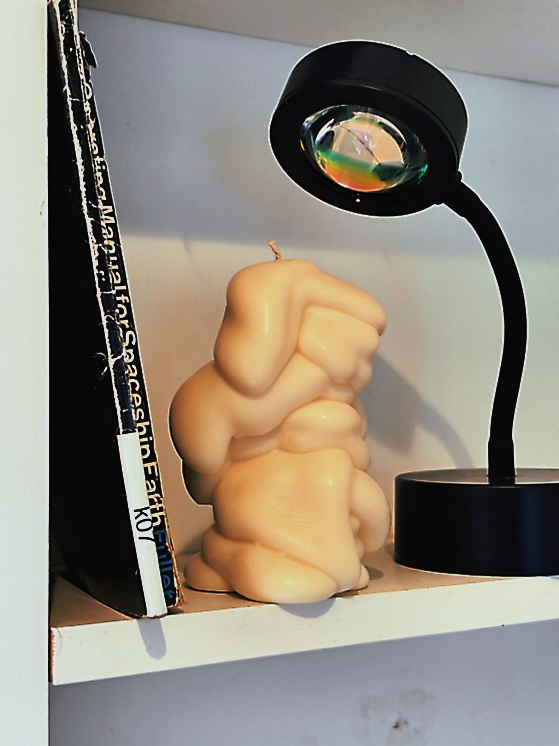White post objects drip drape textured abstract candle on a wooden surface sits on a white shelf next to a black desk lamp and a black book.