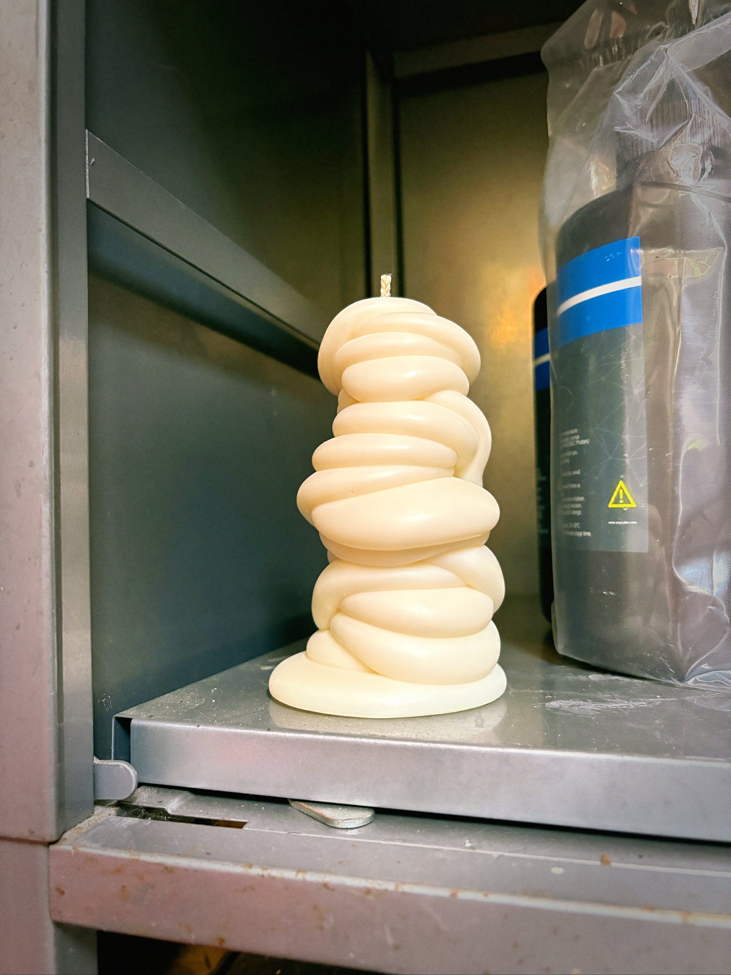 post objects sculptural drip shaped cream-colored candle sits on a stainless steel shelf inside a dark cabinet, next to a dark container wrapped in plastic.