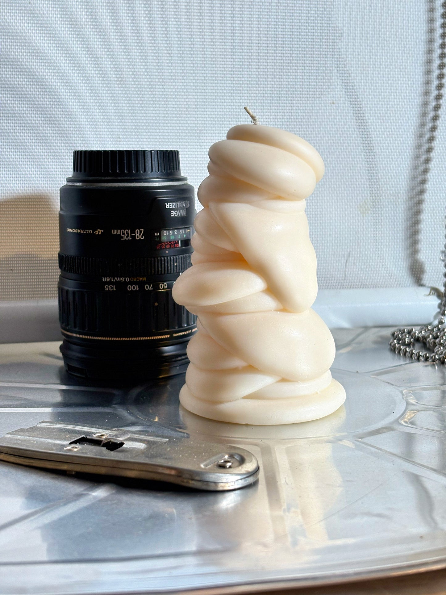 post objects flow series drip art sculptural candle in a warm cream color, styled on a reflective silver tray next to a black camera lens 
