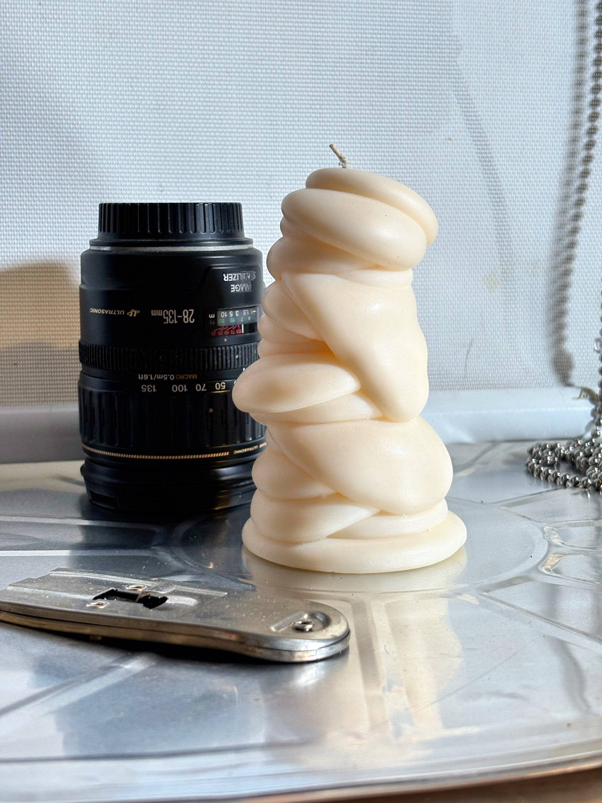 post objects flow series drip art sculptural candle in a warm cream color, styled on a reflective silver tray next to a black camera lens 
