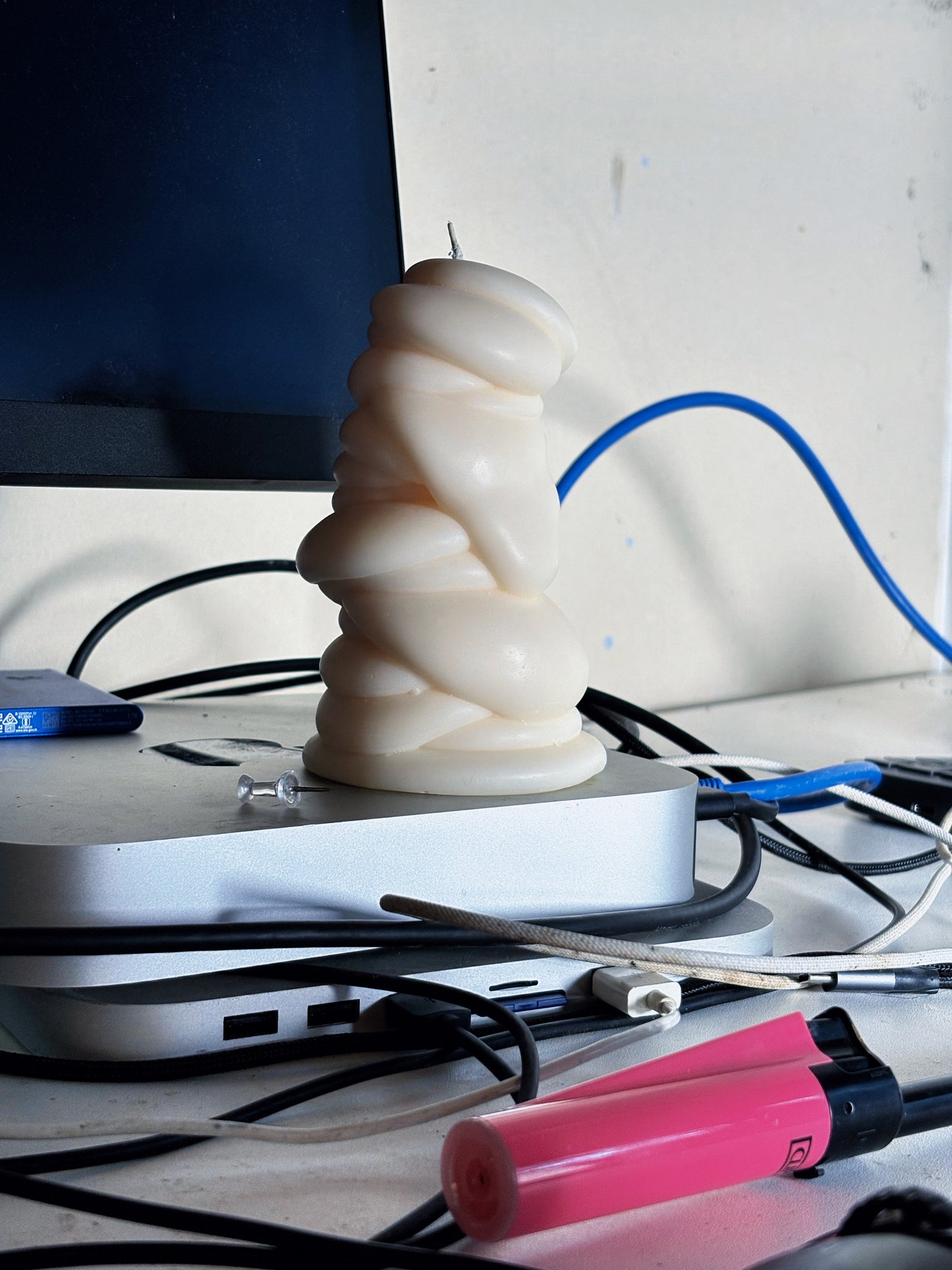 post objects white colored drip sculptural candle sits on top of a silver electronic mac mini device surrounded by tangled black and blue cables on a desk.