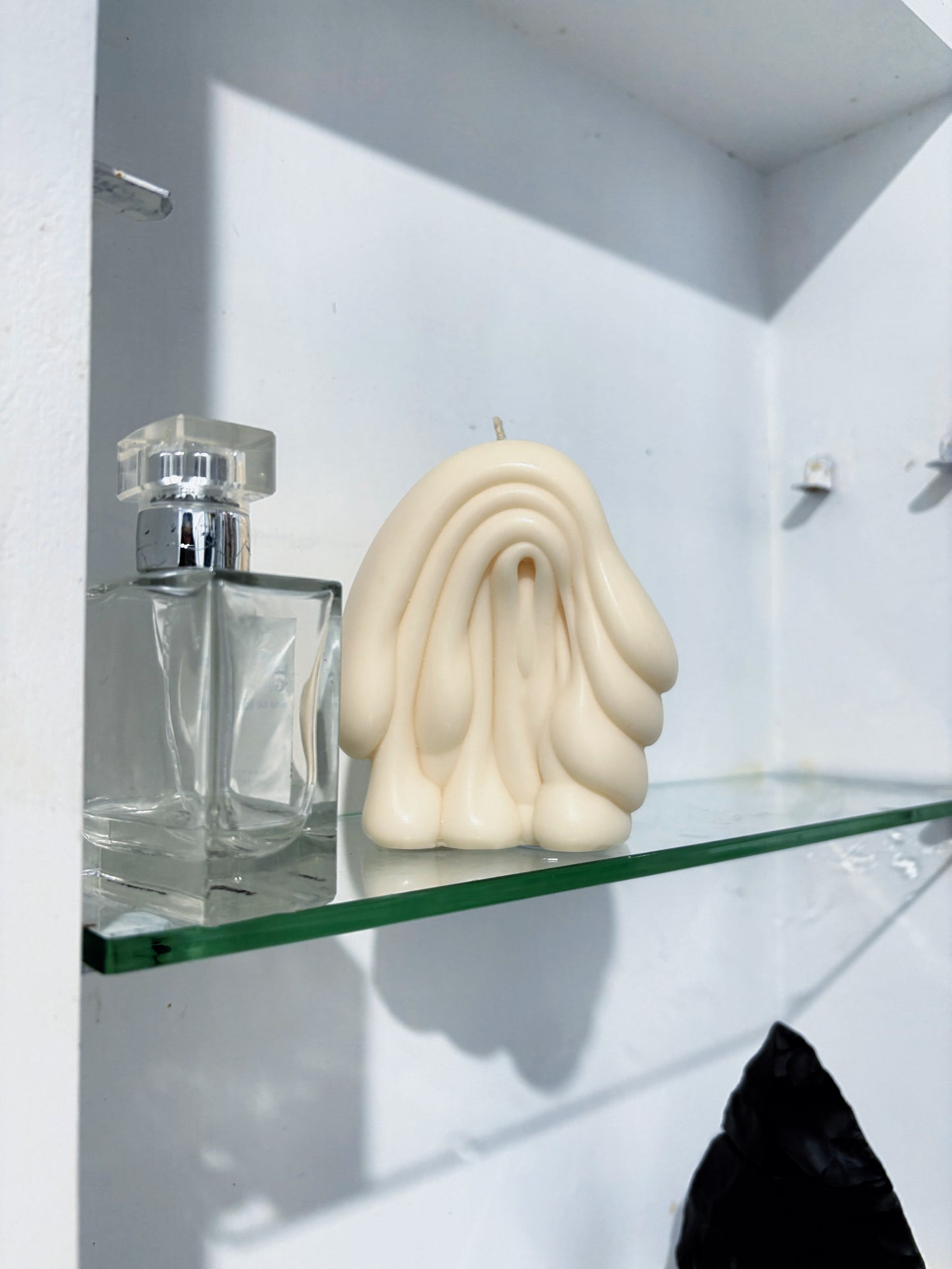 A cream-colored post objects, sculptural hand-shaped candle sits on a clear glass shelf next to a clear glass perfume bottle against a white wall.