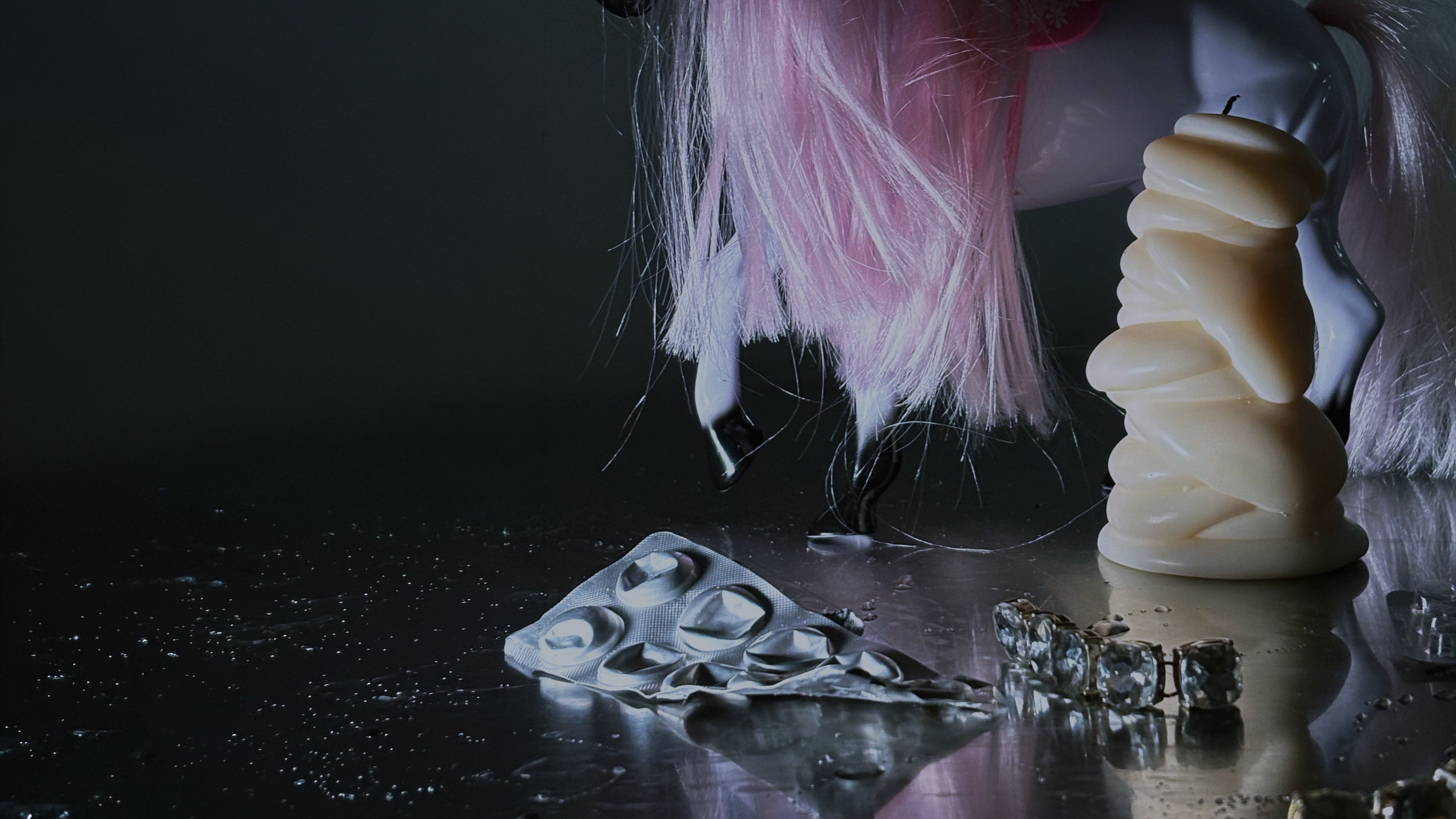 A Post Objects cream-colored sculptural candle on a dark reflective surface with scattered metallic pieces against a dark background.